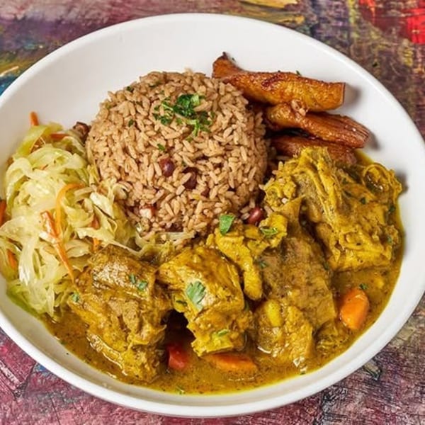 Jamaican curry chicken with rice and peas, fried plantains, and steamed cabbage in a white bowl.