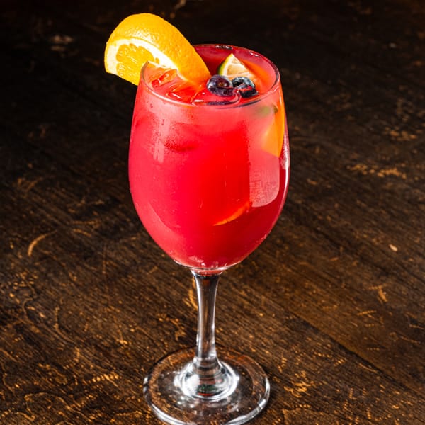 A vibrant red cocktail in a wine glass, garnished with orange, lime, and blueberries on a dark wooden table.