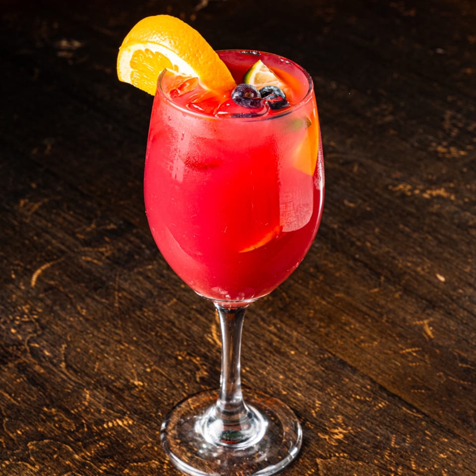 A vibrant red cocktail in a wine glass, garnished with orange, lime, and blueberries on a dark wooden table.