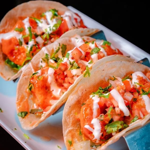Three vibrant Jamaican tacos with red sauce, white crema, and fresh herbs on a white platter.