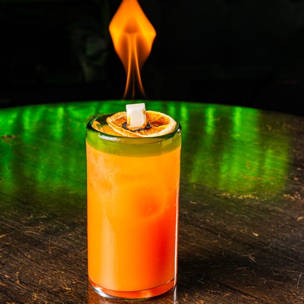 A vibrant orange cocktail with a flaming sugar cube and dried orange slice on a dark table.