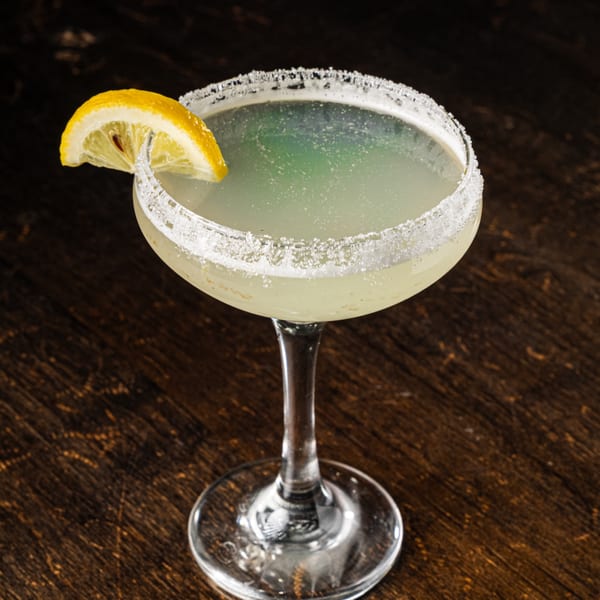 A light green cocktail in a sugar-rimmed coupe glass, garnished with a lemon slice, on a dark wooden surface.